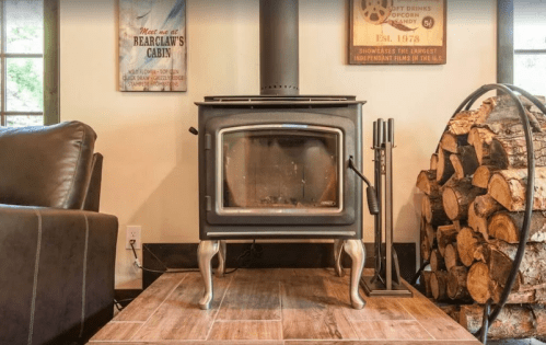A cozy living space featuring a black wood stove, stacked logs, and a wooden floor, with windows in the background.