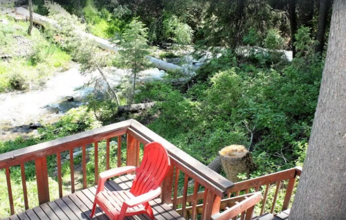 A red chair on a wooden deck overlooks a lush green area with a flowing stream and trees.
