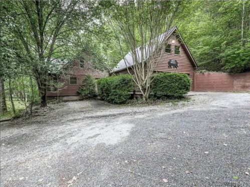 A rustic cabin surrounded by trees, featuring a gravel driveway and a wooden fence in a serene, wooded setting.
