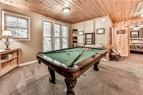 A cozy game room featuring a green pool table, wooden walls, and a lamp beside a shelf. Natural light from windows.