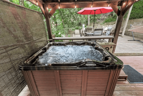 A wooden hot tub under a gazebo, surrounded by greenery and outdoor furniture, with a red umbrella in the background.