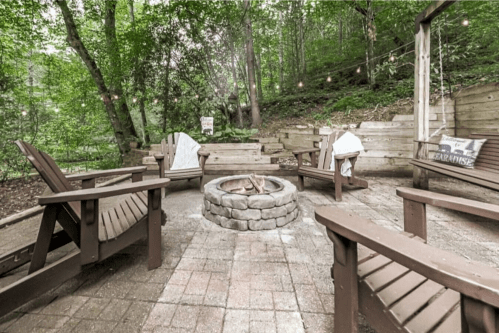 A cozy outdoor fire pit area surrounded by wooden chairs and trees, with string lights overhead.