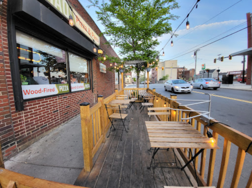Outdoor seating area of a pizza restaurant with wooden tables, string lights, and a view of the street.