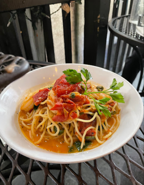 A bowl of pasta topped with shrimp, tomatoes, and herbs, served on a table with a black metal design.