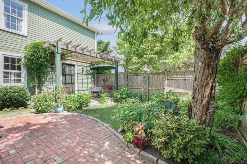 A lush garden with a brick pathway, green plants, and a shaded seating area under a pergola beside a light green house.