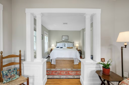 Cozy bedroom with a white bed, wooden floors, and a view through a decorative archway. A chair and lamp are nearby.