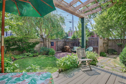 A sunny backyard with a wooden deck, green plants, a pergola, and a flag hanging on the fence.