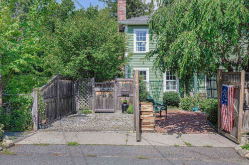 A green house with a wooden fence, gravel pathway, and an American flag hanging on the gate, surrounded by greenery.