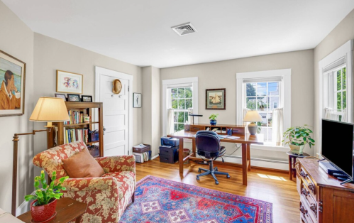 Bright home office with a desk, chair, and cozy seating area, featuring large windows and decorative plants.