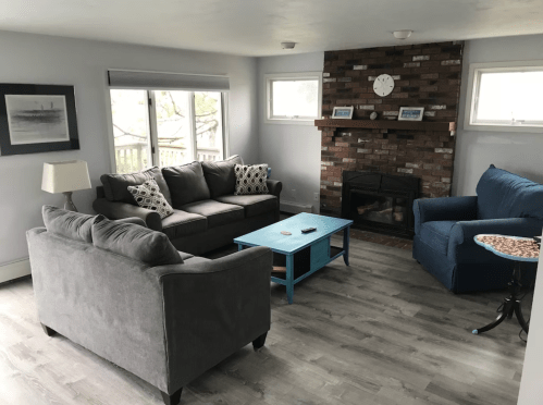 Cozy living room with a gray sofa, blue armchair, brick fireplace, and a light blue coffee table. Natural light from windows.