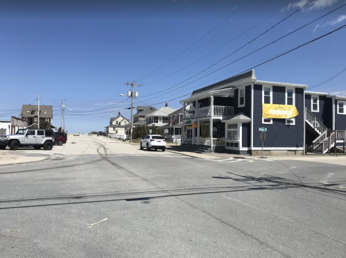 A coastal street scene with a blue building, parked cars, and power lines under a clear blue sky.