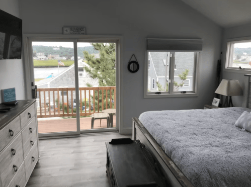 Cozy bedroom with a gray bedspread, dresser, and large windows overlooking a balcony and scenic view.