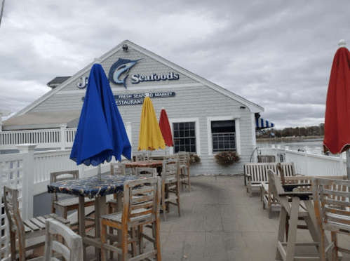 Exterior of a seafood restaurant with colorful umbrellas on a patio, overlooking a body of water under a cloudy sky.