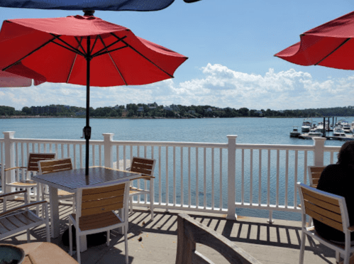 A waterfront view with red umbrellas over tables, boats in the distance, and a clear blue sky.