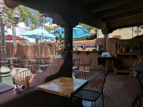 Cozy outdoor café with rustic decor, tables, and blue umbrellas, surrounded by adobe walls and greenery.
