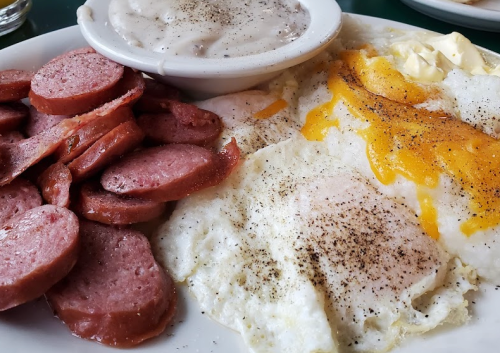 A plate of breakfast featuring scrambled eggs with cheese, grits, sausage slices, and a side of gravy.
