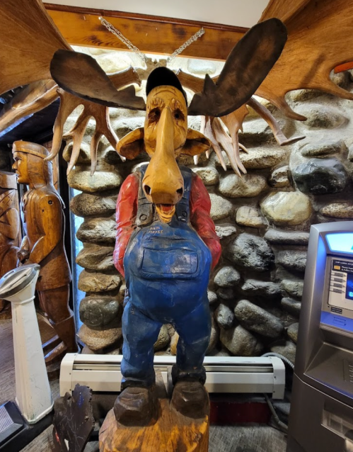 A wooden moose statue wearing overalls and a cap, standing in front of a stone wall.