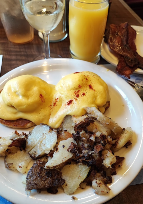 A plate of eggs Benedict with hollandaise sauce, crispy potatoes, a glass of orange juice, and a glass of white wine.