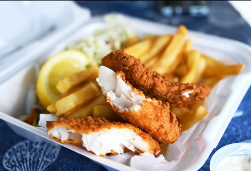 A takeout container with crispy fried fish, golden fries, a lemon wedge, and a side of coleslaw.