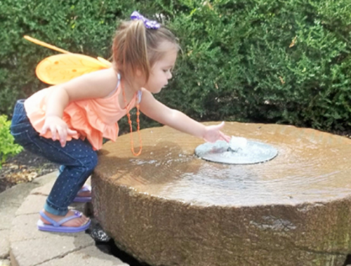 A young girl with butterfly wings reaches into a water fountain, playfully touching the water's surface.