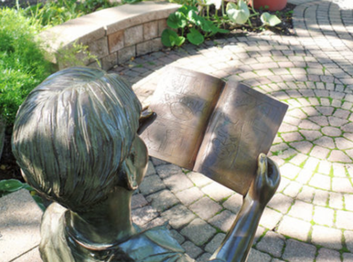 A bronze sculpture of a child reading a book in a garden setting, surrounded by greenery and stone pathways.