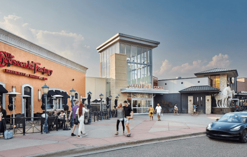 Exterior view of a shopping center featuring The Cheesecake Factory and a modern building, with people walking and cars passing.