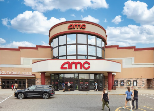 Exterior of an AMC theater with a large sign, people entering, and a car parked in front under a blue sky.