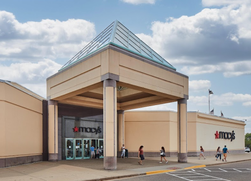 Exterior view of a Macy's store with a glass pyramid roof and shoppers entering on a cloudy day.