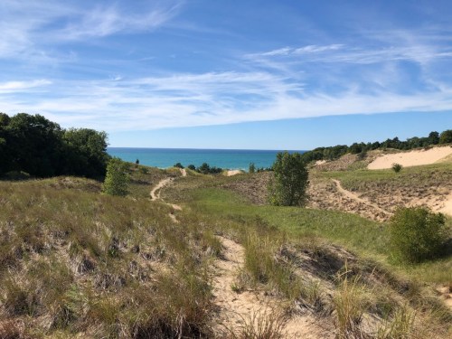 A scenic view of grassy dunes leading to a calm blue lake under a clear sky.
