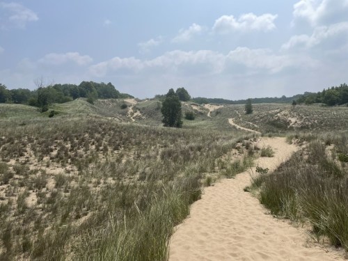 A sandy path winds through grassy dunes under a partly cloudy sky, surrounded by lush greenery.