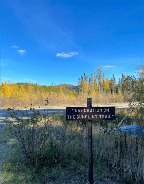 A sign reading "Use caution on the Gunflint Trail" stands in a scenic landscape with trees and a clear blue sky.