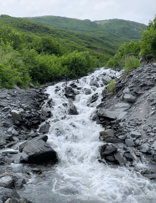 A rocky stream flows through a lush green landscape, surrounded by hills and vegetation.