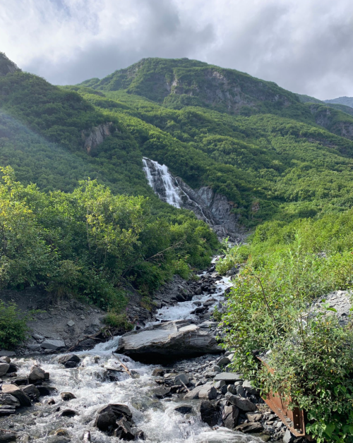 A lush green mountain landscape with a waterfall cascading down rocky terrain, surrounded by dense foliage.
