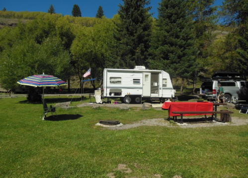 A campsite featuring a white trailer, picnic table, umbrella, and a parked vehicle surrounded by trees and grass.