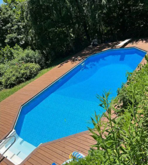 A clear blue swimming pool surrounded by wooden decking and lush greenery.