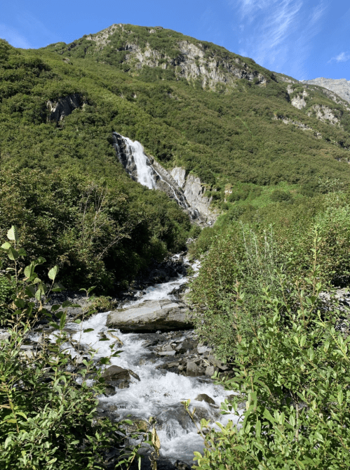 A cascading waterfall flows down a rocky mountain, surrounded by lush greenery and a clear blue sky.