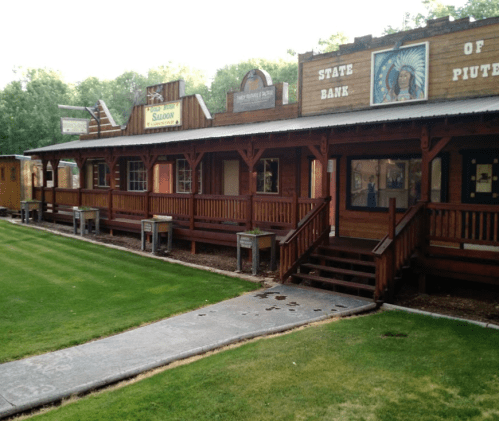 A western-style building with wooden facades, featuring a saloon and a state bank, set in a grassy area.