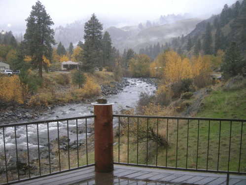A misty river flows through a valley surrounded by autumn trees and mountains, viewed from a wooden deck.