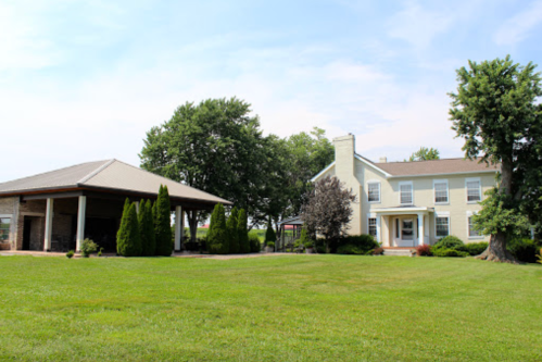 A large, two-story house with a porch, surrounded by green grass and trees, next to a covered outdoor structure.