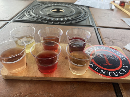 A wooden flight board with eight small cups of various wines, alongside a logo for a Kentucky winery.
