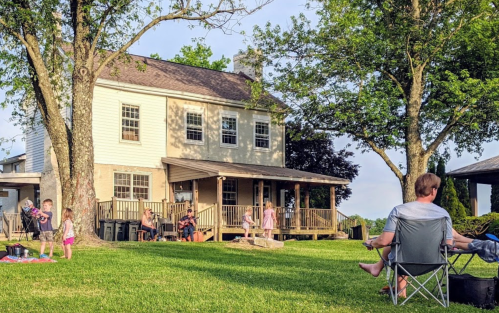 A family gathers outside a two-story house, enjoying a sunny day on the lawn with children playing and adults relaxing.
