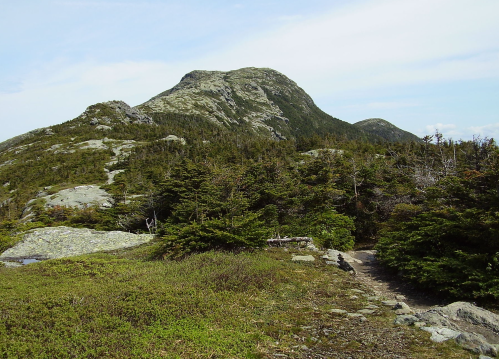 A rocky mountain rises in the background, surrounded by lush greenery and a winding trail in the foreground.