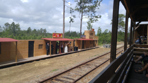 A view of a western-style train station with wooden buildings and a railway track, surrounded by trees and cloudy skies.