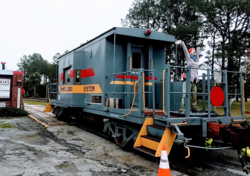 A blue caboose on a railway track, with a person standing on it, surrounded by trees and a traffic cone nearby.
