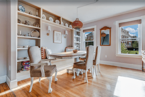 Bright dining room with a wooden table, four chairs, and a shelf displaying dishes, featuring large windows and hardwood floors.