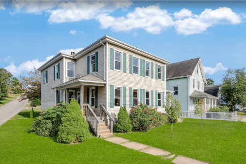 A two-story house with green shutters, surrounded by a lawn and trees, under a blue sky with fluffy clouds.