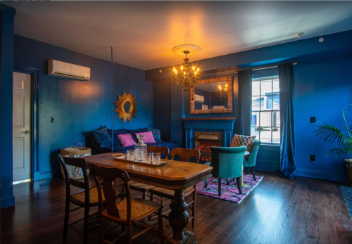 Cozy living room with blue walls, a chandelier, a fireplace, and a dining table surrounded by colorful chairs.