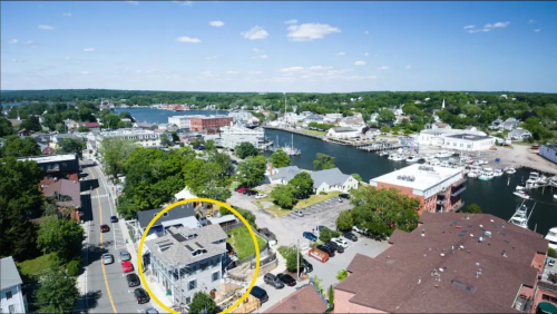 Aerial view of a coastal town with buildings, a marina, and greenery, highlighting a specific structure in a yellow circle.