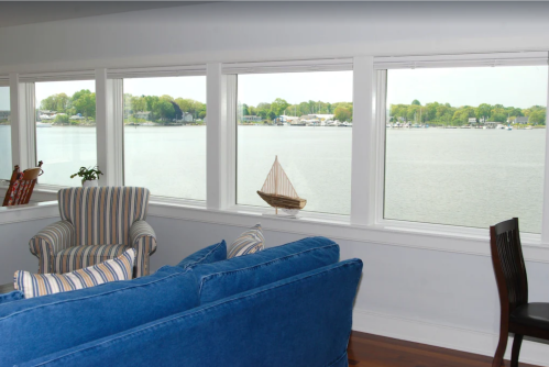 Cozy living room with blue couch and striped chair, featuring large windows overlooking a serene lake and sailboat.