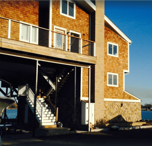 A modern two-story house with wooden siding and a stone base, featuring a staircase and a view of the water.